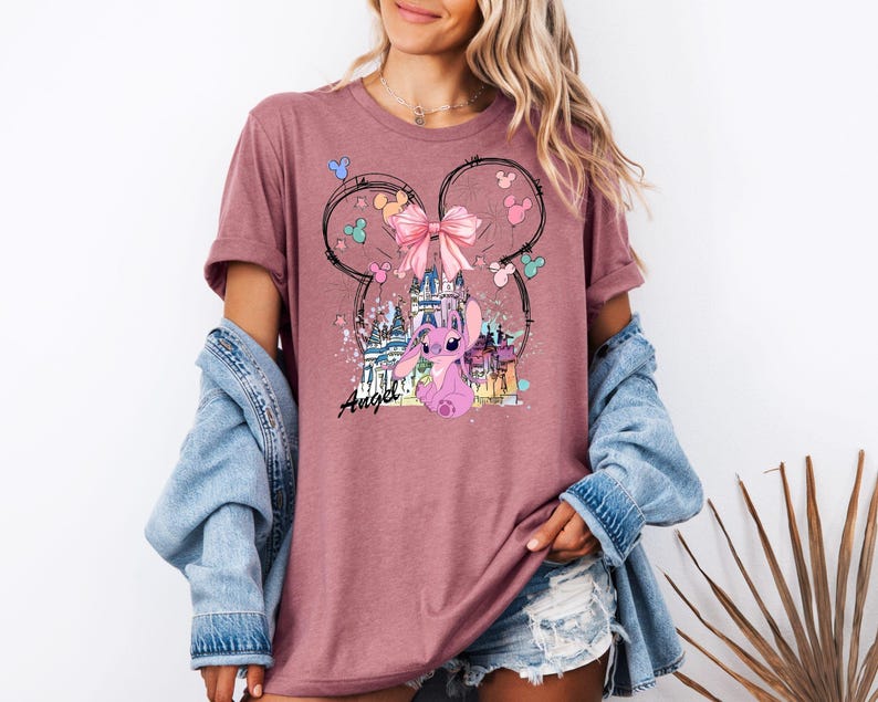 May include: A mauve t-shirt featuring a graphic of a pink cartoon character, a castle, and a pink bow. The design is framed by a Mickey Mouse ear outline with colorful accents. The word "Angel" is written in cursive.