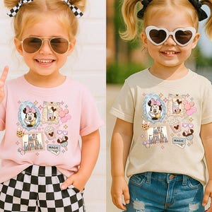 Pu&ograve; includere: Due bambini che indossano magliette con una grafica di Minnie Mouse. La maglietta rosa presenta un personaggio dei cartoni animati di Minnie Mouse in una cornice blu, un castello e altre immagini a tema. La maglietta beige ha un design simile. Entrambe le magliette sono a maniche corte.