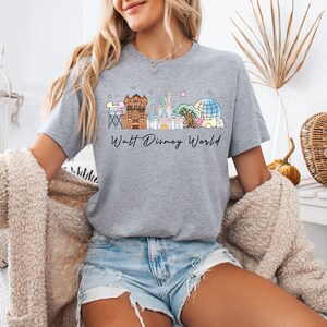 May include: A heather gray short-sleeve t-shirt featuring a colorful graphic of various theme park landmarks and the text Walt Disney World in black script. The design includes pastel-colored illustrations of a water tower, castle, and dome.