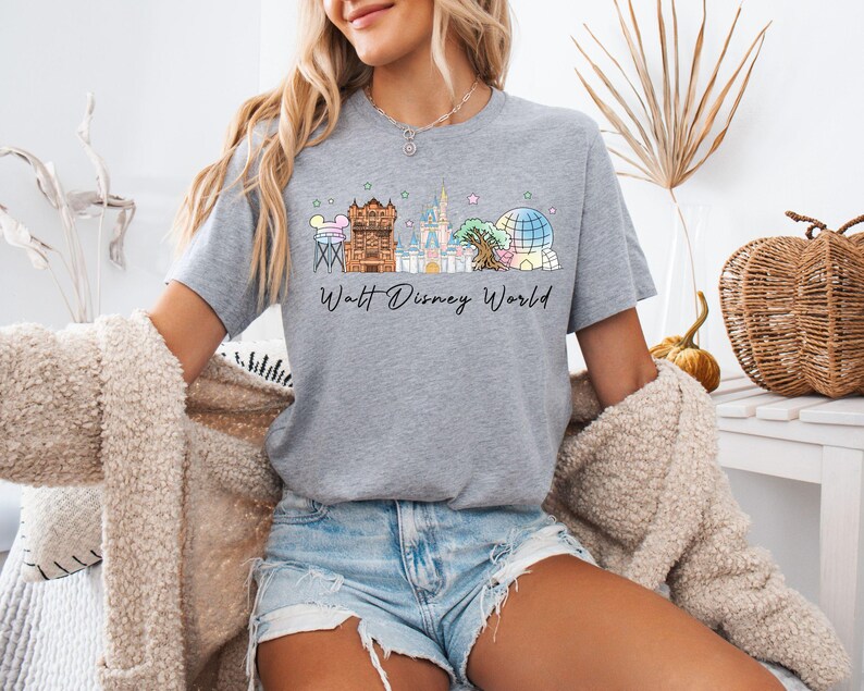 May include: A heather gray short-sleeve t-shirt featuring a colorful graphic of various theme park landmarks and the text Walt Disney World in black script. The design includes pastel-colored illustrations of a water tower, castle, and dome.