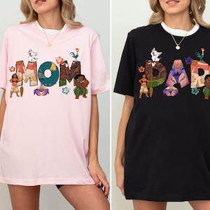 K&ouml;nnte beinhalten: Zwei T-Shirts, eines rosa und eines schwarz, mit einem grafischen Design, das die W&ouml;rter "MOM" und "DAD" in einer stilisierten Schrift zeigt. Die Buchstaben sind mit Figuren aus dem Animationsfilm Vaiana verziert, darunter Maui, Pua und Hei Hei.