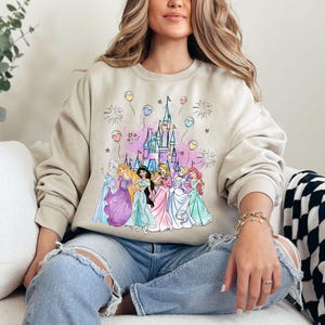 Puede incluir: Una sudadera de color beige claro con un estampado gr&aacute;fico de princesas de Disney y el castillo de Cenicienta. Las princesas llevan vestidos de color pastel y est&aacute;n rodeadas de fuegos artificiales y globos. El texto "Let's Go!" est&aacute; impreso en el castillo.