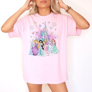 Puede incluir: Camiseta rosa con un gr&aacute;fico de Cenicienta, Blancanieves, Ariel, Bella, Jasmine, Mulan, Tiana, Rapunzel y Pocahontas de pie frente al castillo de Cenicienta con fuegos artificiales y globos.