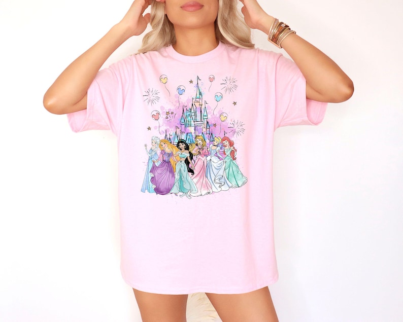 Puede incluir: Camiseta rosa con un gr&aacute;fico de Cenicienta, Blancanieves, Ariel, Bella, Jasmine, Mulan, Tiana, Rapunzel y Pocahontas de pie frente al castillo de Cenicienta con fuegos artificiales y globos.