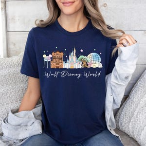 May include: A navy blue short-sleeve t-shirt featuring a graphic of the Walt Disney World skyline, including the castle, water tower, and park icons in pastel colors, with the text Walt Disney World printed in white cursive script below the design.