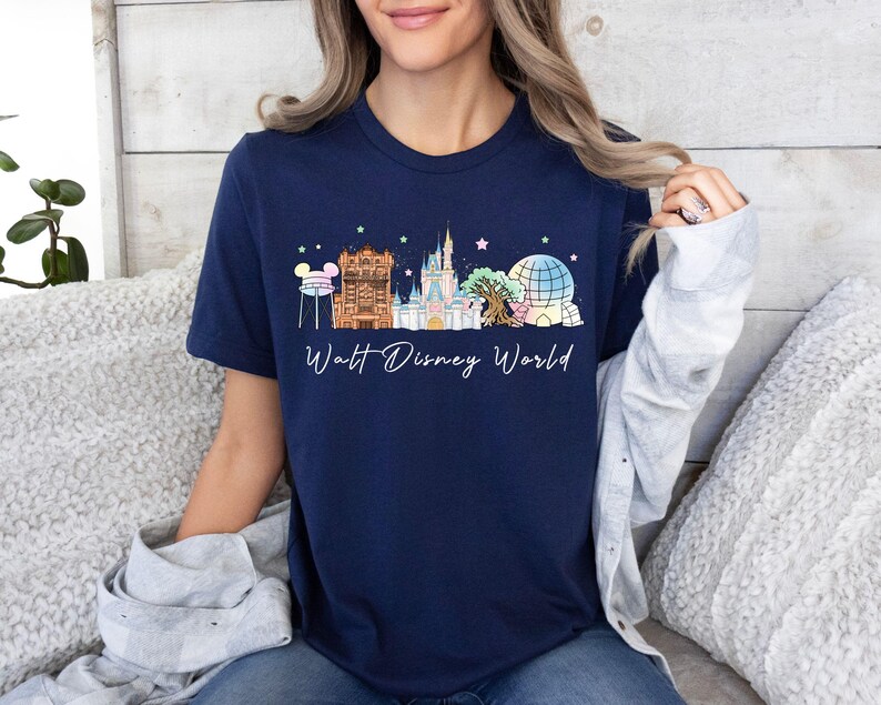 May include: A navy blue short-sleeve t-shirt featuring a graphic of the Walt Disney World skyline, including the castle, water tower, and park icons in pastel colors, with the text Walt Disney World printed in white cursive script below the design.