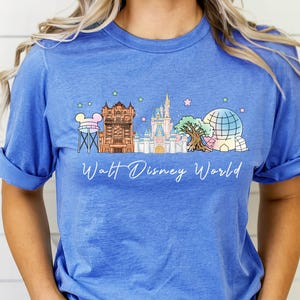 May include: A blue short-sleeve t-shirt featuring a graphic print of the Walt Disney World skyline, including the water tower, Hollywood Tower Hotel, Cinderella Castle, Tree of Life, and Spaceship Earth, with small pastel stars above.