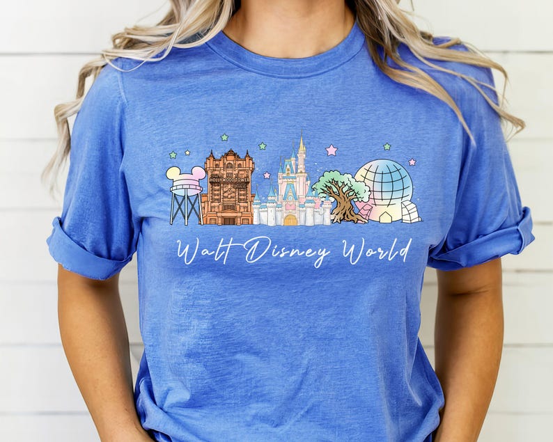 May include: A blue short-sleeve t-shirt featuring a graphic print of the Walt Disney World skyline, including the water tower, Hollywood Tower Hotel, Cinderella Castle, Tree of Life, and Spaceship Earth, with small pastel stars above.