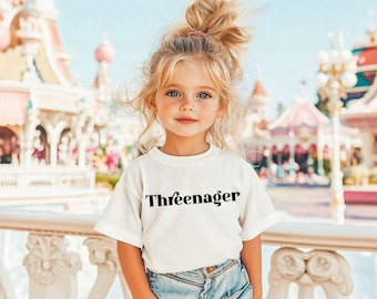May include: A young child wearing a white t-shirt with the word "Threenager" printed in black. The child has blonde hair and is standing in front of a colorful background.