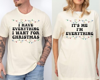 May include: Two cream-coloured t-shirts with black text and colourful Christmas lights. One shirt reads "I HAVE EVERYTHING I WANT FOR CHRISTMAS." The other says "IT'S ME I'M EVERYTHING." Both shirts have a relaxed fit.