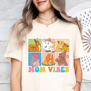 Può includere: T-shirt color crema con una grafica colorata di personaggi Disney e dei loro bambini, tra cui Bambi, Mrs. Potts, Simba, Dumbo, Lady e Roo. Le parole "MOM VIBES" sono scritte in un font retrò.