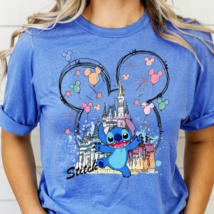 May include: Blue t-shirt featuring a graphic of Stitch from Lilo & Stitch in front of a castle, fireworks, and Mickey Mouse ears. The word "Stitch" is written in black script below the character.