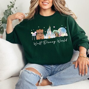 May include: A dark green crewneck sweatshirt featuring a graphic print of the Walt Disney World skyline, including the castle, water tower, and park icons, with the text Walt Disney World written in white cursive script below the illustration.