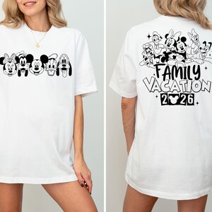 Peut inclure: T-shirt blanc avec un motif noir et blanc de personnages Disney, dont Minnie Mouse, Mickey Mouse et Pluto. Le dos du t-shirt affiche le texte "FAMILY VACATION 2026" avec des illustrations de personnages.