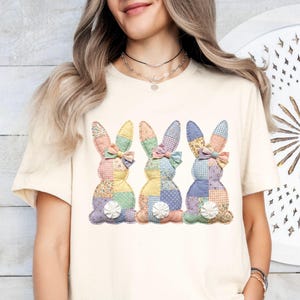 May include: Cream-colored t-shirt featuring three patchwork bunny designs. Each bunny has a different color scheme, including pink, yellow, blue, and green, with a bow and a white flower detail. The bunnies are arranged in a row.