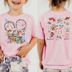 Op de afbeelding: Een lichtroze T-shirt met een Disney-thema. De voorkant toont een Mickey Mouse oor-omtrek met vuurwerk en Disney-personages. De achterkant toont karakterportretten en namen, waaronder Woody en Buzz Lightyear.