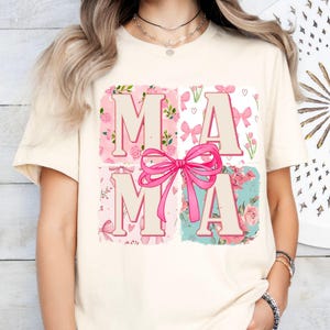 Puede incluir: Una camiseta de color crema con un diseño floral rosa y blanco. El diseño presenta la palabra "MAMA" con un lazo rosa en el centro.