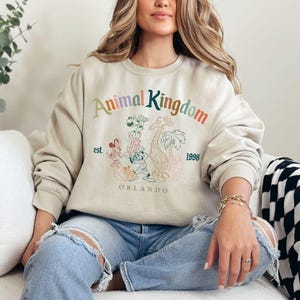 K&ouml;nnte beinhalten: Beiges Sweatshirt mit Rundhalsausschnitt und dem Text "Animal Kingdom" in Regenbogenfarben, Cartoon-Tierillustrationen und dem Text "est. 1998 Orlando". Das Sweatshirt hat einen l&auml;ssigen Stil.