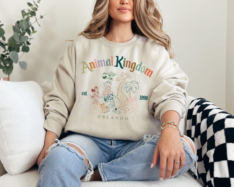 K&ouml;nnte beinhalten: Beiges Sweatshirt mit Rundhalsausschnitt und dem Text "Animal Kingdom" in Regenbogenfarben, Cartoon-Tierillustrationen und dem Text "est. 1998 Orlando". Das Sweatshirt hat einen l&auml;ssigen Stil.