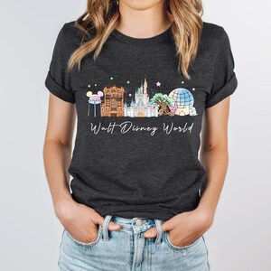 May include: A dark charcoal gray short-sleeve t-shirt featuring a graphic of the Walt Disney World skyline, including the castle, iconic park structures, and colorful stars. The text Walt Disney World is printed in white script below the graphic.