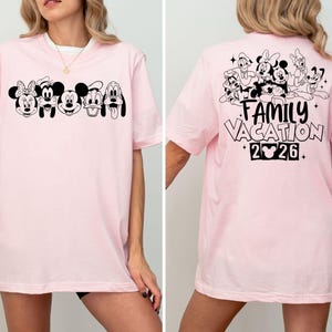 Peut inclure: T-shirt rose clair avec des visages de personnages de dessins anim&eacute;s en noir et blanc sur le devant. Le dos du t-shirt affiche un groupe de personnages de dessins anim&eacute;s et le texte "FAMILY VACATION 2026".