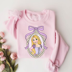 May include: Pink sweatshirt with a purple bow and a picture of Rapunzel in a pearl frame.