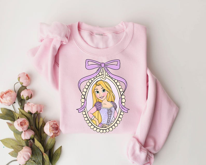 May include: Pink sweatshirt with a purple bow and a picture of Rapunzel in a pearl frame.