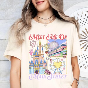 以下が含まれることがあります： クリーム色のTシャツ。カラフルなグラフィックには「Meet Me On Main Street」の文字と、お城、モノレール、ミッキーマウスの観覧車、その他のテーマパークの要素のイラストが描かれています。