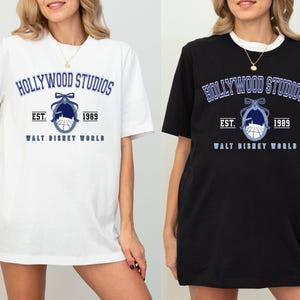 Puede incluir: Camisetas blancas y negras con el texto "HOLLYWOOD STUDIOS" en azul, un gr&aacute;fico a juego y el texto "EST. 1989 WALT DISNEY WORLD". Las camisetas son de manga corta y tienen un corte holgado.