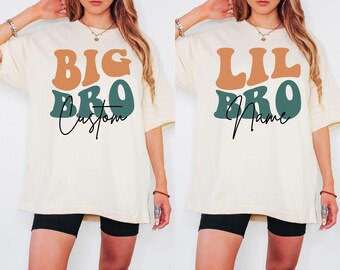 Puede incluir: Dos camisetas blancas con un diseño de fuente retro. Una camiseta dice "BIG BRO Custom" y la otra dice "LIL BRO Name".