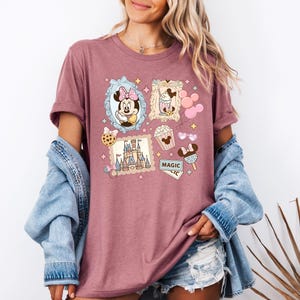 Pu&ograve; includere: T-shirt color malva con stampa collage a tema Disney. Il design include immagini di Minnie Mouse, un castello, popcorn e gelato. La parola "MAGIC" &egrave; visibile anche sulla maglietta.