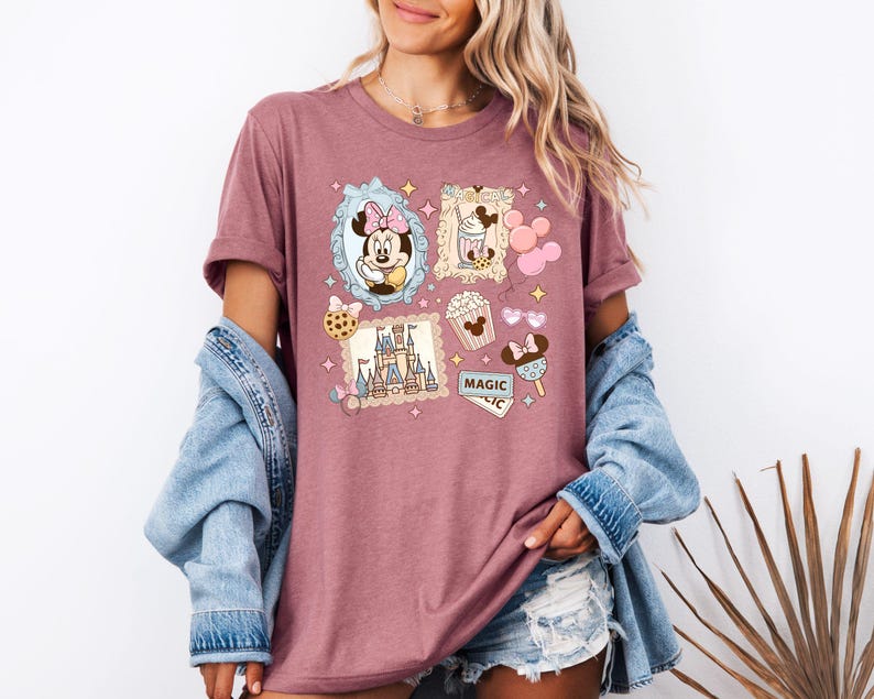 Pu&ograve; includere: T-shirt color malva con stampa collage a tema Disney. Il design include immagini di Minnie Mouse, un castello, popcorn e gelato. La parola "MAGIC" &egrave; visibile anche sulla maglietta.