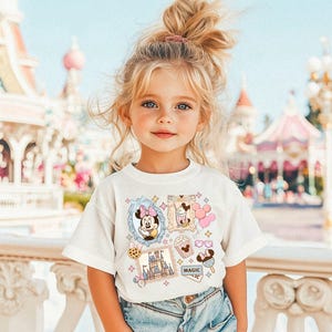 Pu&ograve; includere: Una bambina indossa una maglietta bianca con grafiche colorate a tema Disney. La maglietta presenta immagini di Minnie Mouse, un castello, gelato e altre icone correlate. La bambina &egrave; in piedi di fronte a un parco a tema.