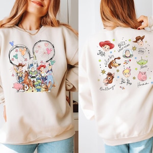 Op de afbeelding: Beige sweatshirt met een kleurrijk Toy Story-ontwerp. De voorkant toont Mickey Mouse oren met vuurwerk en ballonnen. De achterkant toont karakterportretten en namen zoals Buzz Lightyear en Woody.