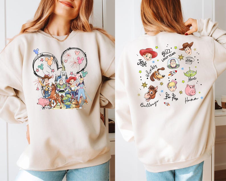 Op de afbeelding: Beige sweatshirt met een kleurrijk Toy Story-ontwerp. De voorkant toont Mickey Mouse oren met vuurwerk en ballonnen. De achterkant toont karakterportretten en namen zoals Buzz Lightyear en Woody.