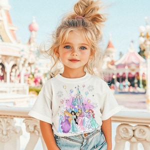 Puede incluir: Una ni&ntilde;a peque&ntilde;a con una camiseta blanca con una ilustraci&oacute;n colorida de princesas Disney y el castillo de Cenicienta. La camiseta tiene una combinaci&oacute;n de colores rosa, morado y azul con detalles de purpurina. La ni&ntilde;a est&aacute; de pie frente a un carrusel rosa y blanco.