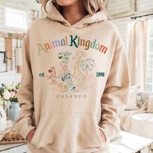 K&ouml;nnte beinhalten: Beiger Kapuzenpullover mit Frontaufdruck. Der Aufdruck zeigt den Text "Animal Kingdom" in verschiedenen Farben, mit "est. 1998" und "Orlando" darunter. Die Grafik enth&auml;lt Zeichentricktiere und eine Giraffe.