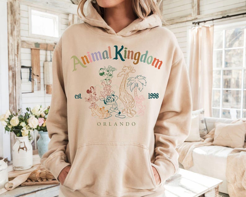 K&ouml;nnte beinhalten: Beiger Kapuzenpullover mit Frontaufdruck. Der Aufdruck zeigt den Text "Animal Kingdom" in verschiedenen Farben, mit "est. 1998" und "Orlando" darunter. Die Grafik enth&auml;lt Zeichentricktiere und eine Giraffe.