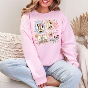 Pu&ograve; includere: Una felpa rosa chiaro con un collage di immagini di cartoni animati. Le immagini includono Minnie Mouse, un castello, gelati e altre grafiche a tema Disney. &Egrave; visibile anche la parola "MAGIC". La felpa ha maniche lunghe e girocollo.