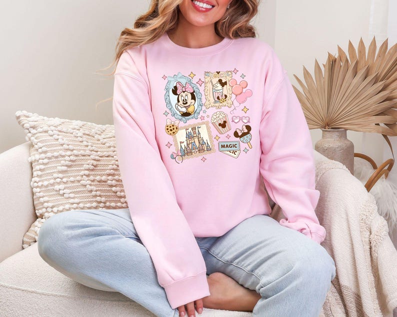 Pu&ograve; includere: Una felpa rosa chiaro con un collage di immagini di cartoni animati. Le immagini includono Minnie Mouse, un castello, gelati e altre grafiche a tema Disney. &Egrave; visibile anche la parola "MAGIC". La felpa ha maniche lunghe e girocollo.