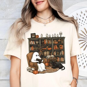 May include: Cream t-shirt with a Halloween graphic. It shows a ghost reading in an armchair before a bookshelf with books, pumpkins, and candles. A black cat and skulls are also included in the design.