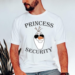 以下が含まれることがあります： サングラスとニンジンの鼻をした漫画のキャラクター、オラフが描かれた白いTシャツ。「PRINCESS SECURITY」の文字がキャラクターの上と下に黒い書体でプリントされています。半袖のTシャツです。