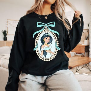 May include: Black sweatshirt with a blue and gold illustration of Princess Jasmine from Disney's Aladdin. The illustration is framed with a gold and white pearl border and a blue bow.