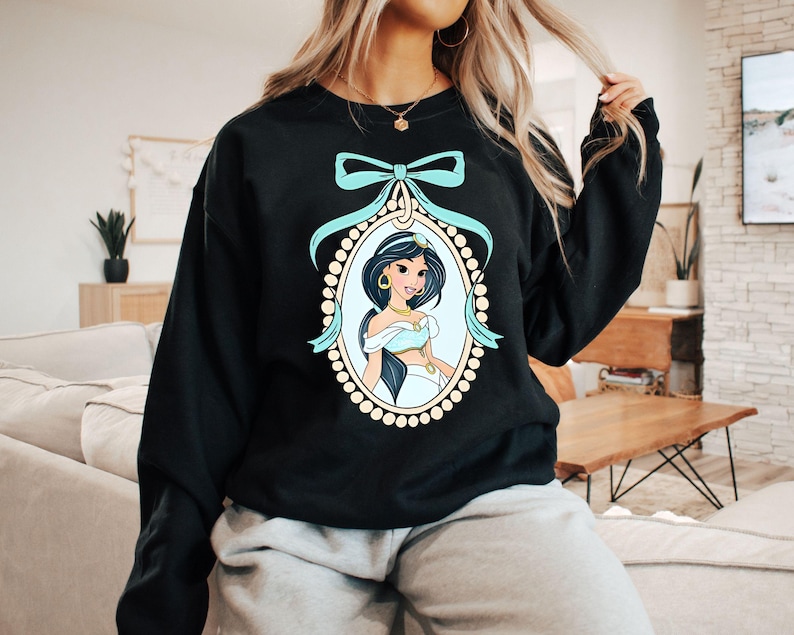 May include: Black sweatshirt with a blue and gold illustration of Princess Jasmine from Disney's Aladdin. The illustration is framed with a gold and white pearl border and a blue bow.