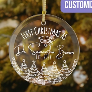 May include: A clear glass Christmas ornament with the text "First Christmas as Dr. Samantha Brown Est. 2024" and a graduation cap and diploma design. The ornament is surrounded by a white outline of pine trees.