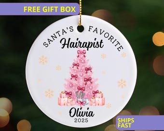May include: A white ceramic Christmas ornament with a pink Christmas tree design. The ornament reads "Santa's Favorite Hairapist" with the name "Olivia" and the year "2025" below the tree. Gold snowflakes and bows decorate the tree.