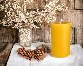 May include: A yellow beeswax candle with a white wick sits on a white linen tablecloth with pine cones. The candle is in front of a vase with white flowers.