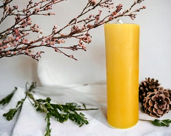 May include: A large, yellow beeswax candle on a white linen tablecloth with a sprig of green foliage and a pine cone.