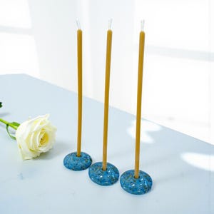 May include: Three yellow taper candles in speckled blue ceramic holders. A white rose lies on the table. The candles are tall and slender, with a simple, elegant design. The holders have a textured, organic appearance.