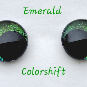 May include: Two round, black-backed, glass eyes with a green and blue iridescent color shift. The eyes are labeled "Emerald" and "Colorshift".